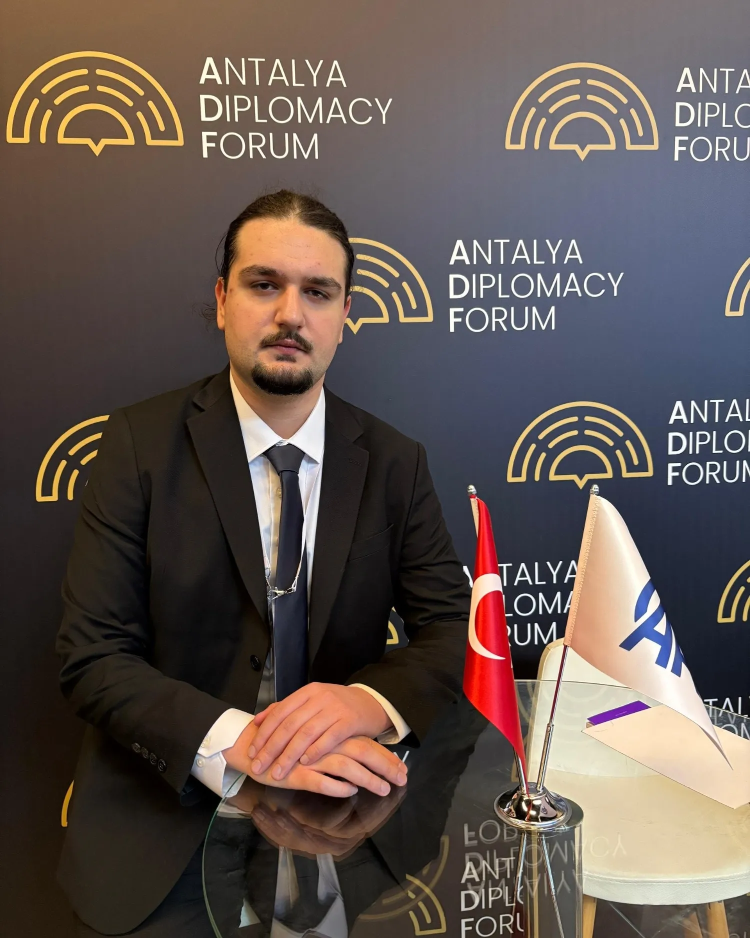 Buğra Öngen, Antalya Diplomasi Forumu’nun logolu panosu önünde bir masada oturuyor; koyu takım elbise ve kravatla, masada bir Türk bayrağı.